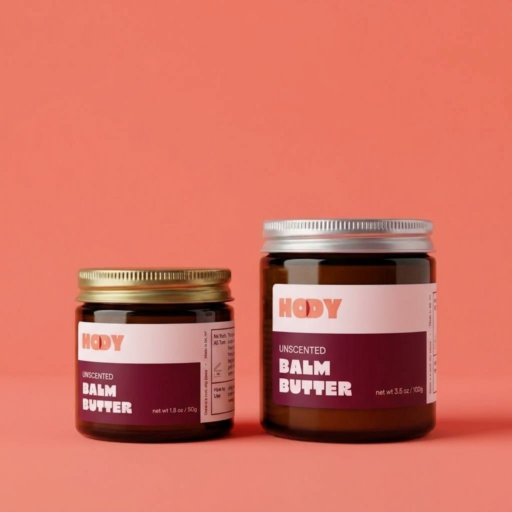 HODY Unscented Balm Butter in small and large amber glass jars on a coral background
