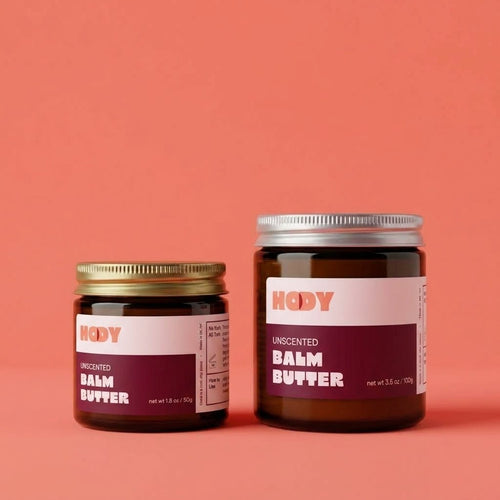 HODY Unscented Balm Butter in small and large amber glass jars on a coral background